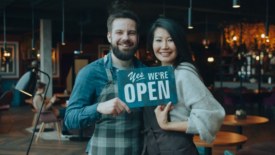 Two cafe owners holding a "We're open" sign inside their small cafe