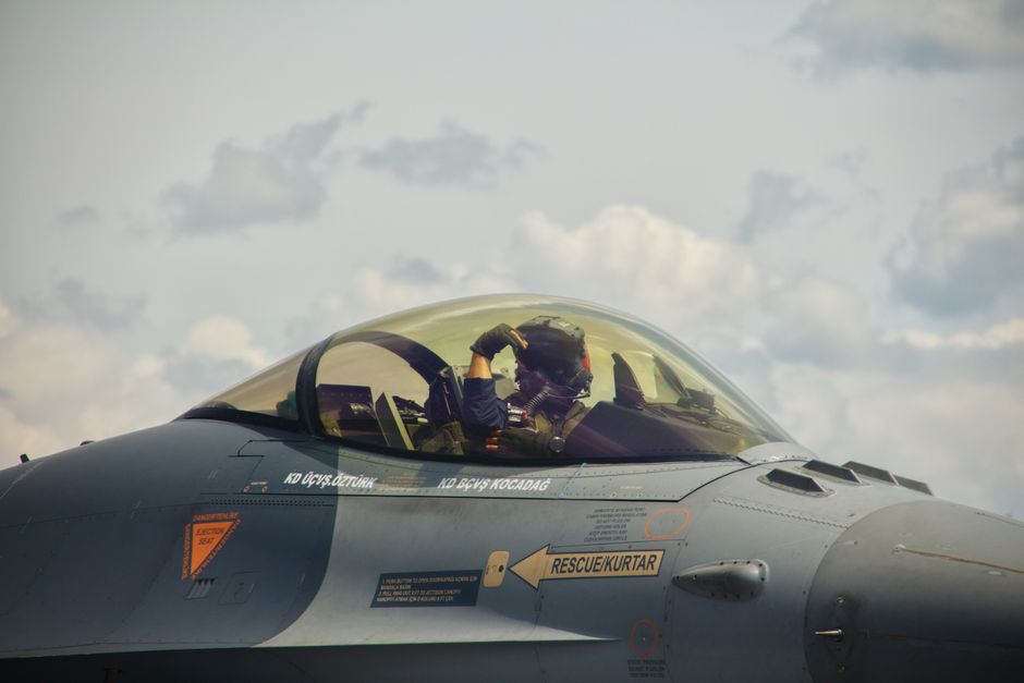 A military pilot sits in the cockpit of a fighter jet, ready for takeoff under a cloudy sky.
