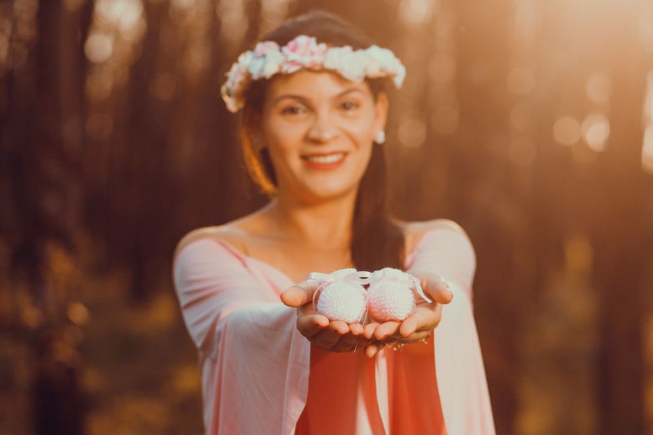 A joyful pregnant woman holds baby shoes, symbolizing new beginnings in a serene outdoor setting.