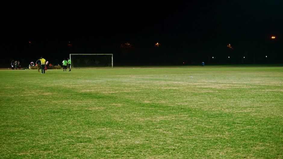 View of a soccer field at night with players and goalposts under floodlights.