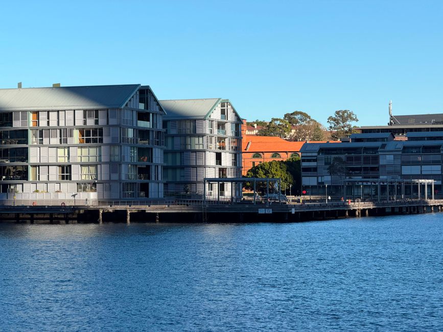 Modern waterfront buildings with glass facades under a clear blue sky, reflecting on the water.