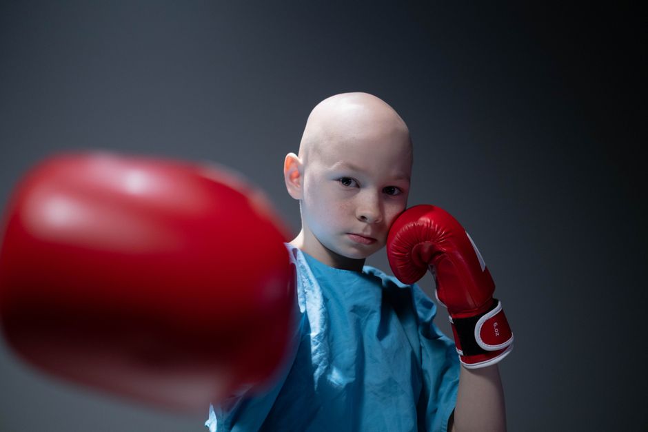 Bald child in boxing stance wearing hospital gown and red gloves, symbolizing strength.