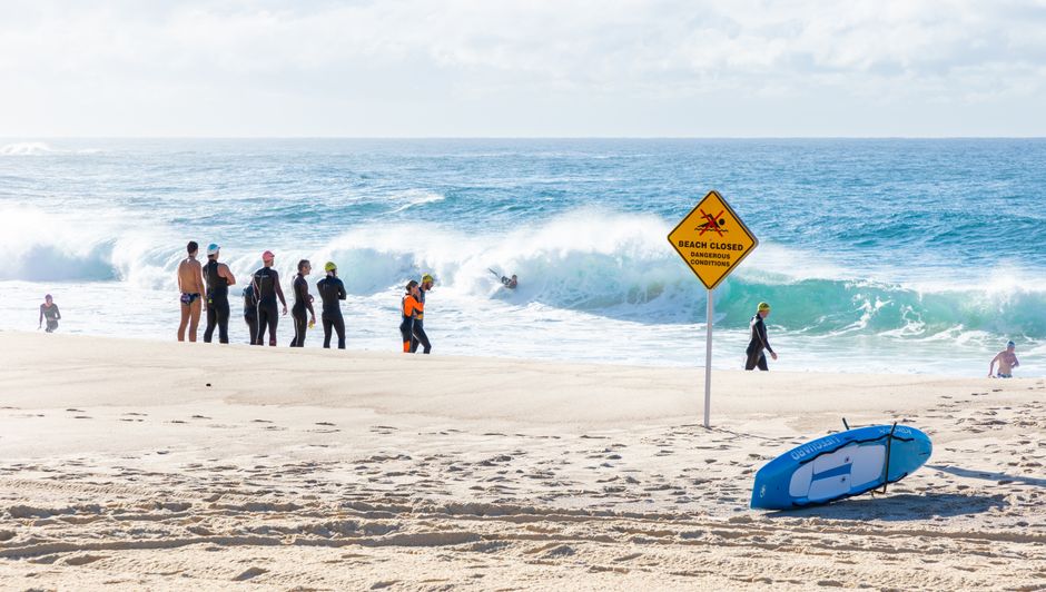 Group of surfers on a beach with a 'Beach Closed' sign and waves in the background.